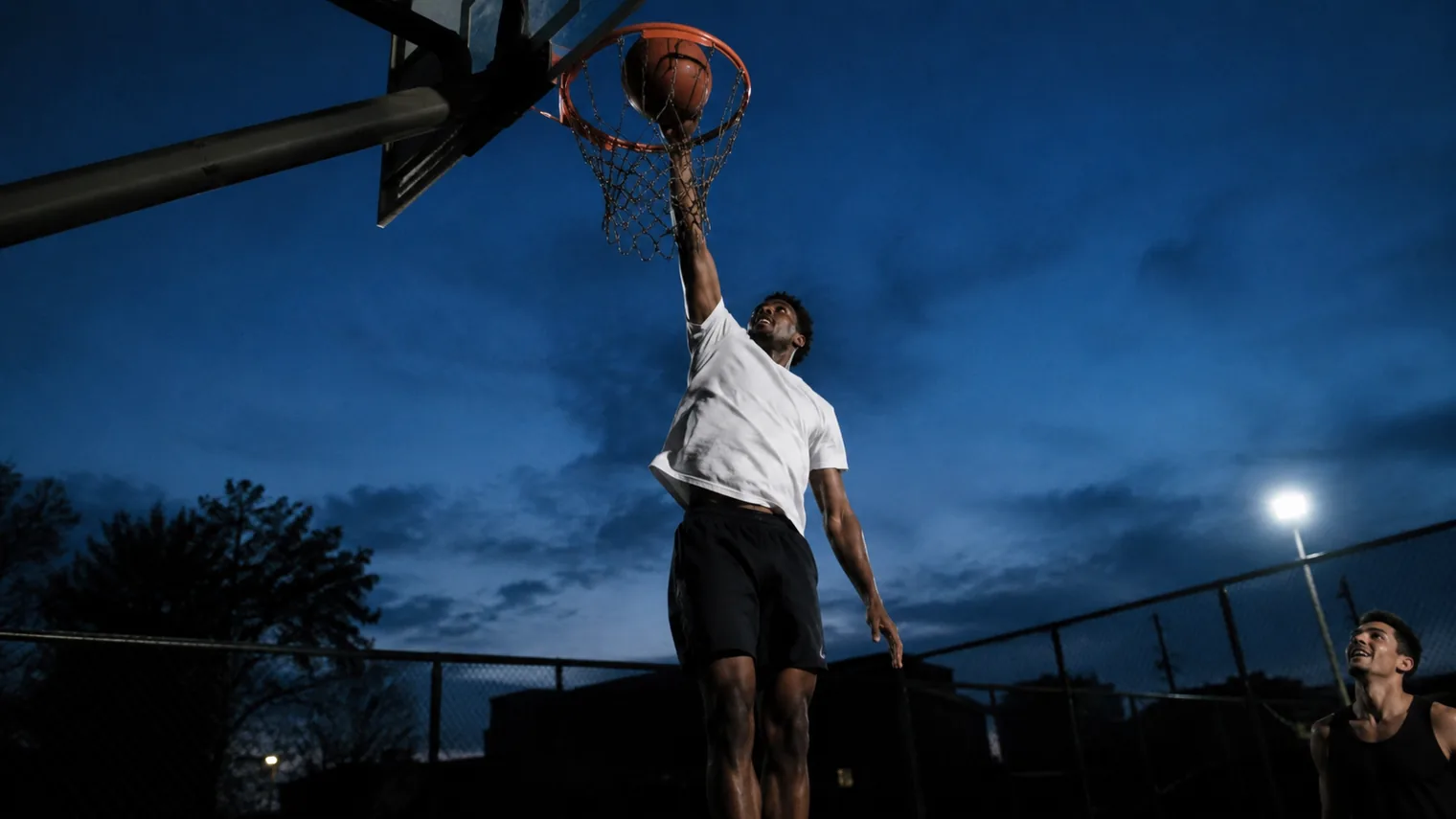 A player at the peak of a one-hand dunk on an outdoor rim at dusk