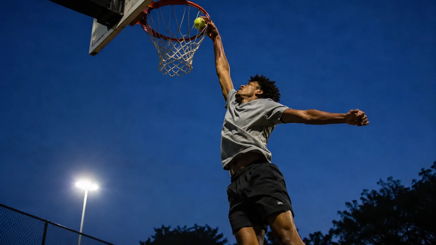 A player at the peak of a jump dunking a tennis ball through an outdoor rim